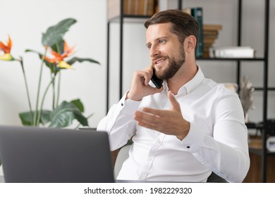 Successful Caucasian Businessman Talking By Smartphone With Somebody While Working Laptop In The Office. Executive Young Businessman Talking On Mobile Phone While Sitting At Work Desk