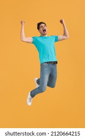 Successful caucasian guy celebrating triumph and jumping in air. Modern young millennial man. Dark-haired handsome male person wearing t-shirt. Isolated on orange background. Studio shoot. Copy space - Shutterstock ID 2120664215