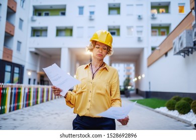 Successful Caucasian Senior Female Architect With Helmet On Head Looking At Documents While Standing Outdoors.