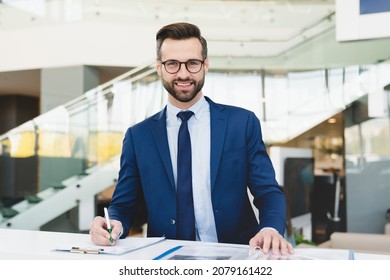 Successful caucasian smiling man shop assistant receptionist in formal attire writing looking at camera while standing at reception desk in hotel car dealer shop
