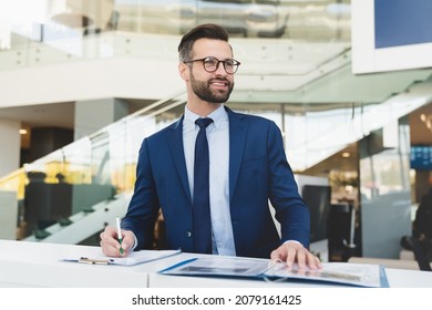 Successful caucasian smiling man shop assistant receptionist in formal attire writing while standing at reception desk in hotel car dealer shop
