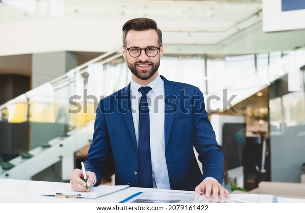 Successful caucasian smiling man shop\
assistant receptionist in formal attire writing looking at camera\
while standing at reception desk in hotel car dealer\
shop