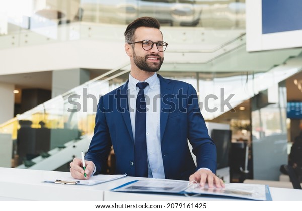 Successful caucasian smiling man shop assistant\
receptionist in formal attire writing while standing at reception\
desk in hotel car dealer\
shop