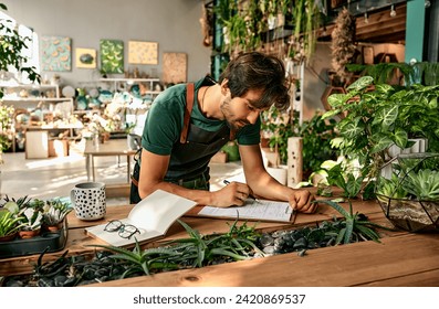 Successful confident business man in green t-shirt and black apron working in greenhouse indoors. Florist writing on clipboard at wooden desk with plants.                             - Powered by Shutterstock