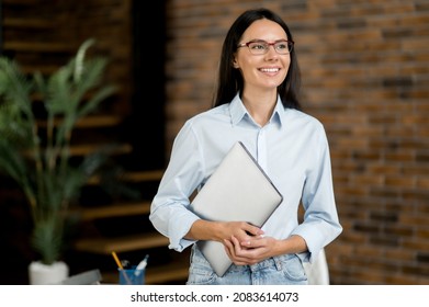 Successful confident pretty young adult caucasian business lady or manager, stand near work desk in office, wearing formal stylish clothes and glasses, holds a laptop, looking away, smiles friendly - Shutterstock ID 2083614073
