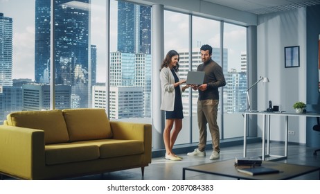 Successful Corporate CEO and Investment Manager Talking, Using Laptop Computer while Standing in Big City Office. Two Successful Diverse People Brainstorm Their e-Commerce Software Investment Strategy