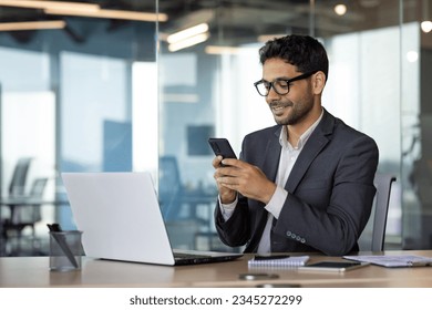 Successful experienced businessman using phone while sitting at workplace, hispanic smiling happy with achievement results holding phone, reading online using app on smartphone. - Shutterstock ID 2345272299