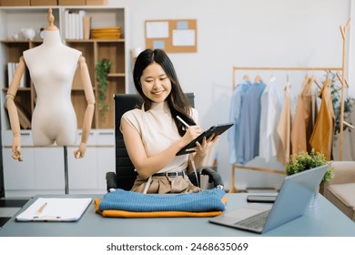 Successful fashion designer. Attractive young asian woman with smile while standing in workshop. Beautiful owner business woman working and holds tablet, laptop and smartphone  - Powered by Shutterstock