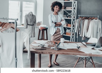 Successful fashion designer. Full length of attractive young African woman keeping arms crossed and looking at camera with smile while standing in workshop 