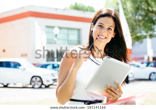 Successful female car sales\
consultant in business trade fair showing blank digital tablet\
screen and approving with thumbs up. Vehicle offer and information\
concept.