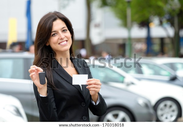 Successful female luxury car sales\
representative showing car key and business card  in automobile\
trade fair. Beautiful brunette saleswoman\
outdoor.