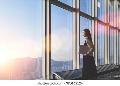 Successful female office worker with net-book is standing in skyscraper interior against big window with city view on background. Proud asian woman architect looking satisfied with completed project