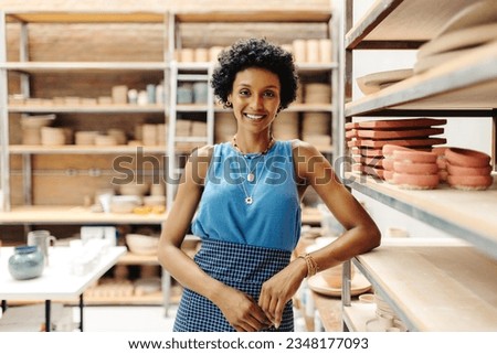 Successful female potter smiling at the camera while standing next to her handmade ceramic products in her shop. Happy young craftswoman running a creative small business.