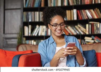 Successful female student with dark skin wearing jean shirt and glasses sharing her success with his friends via social networks after passed exam while sitting in reading hall at comfortable couch - Powered by Shutterstock