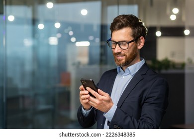 Successful financier investor works inside office at work, businessman in business suit uses telephone near window, man smiles and reads good news online from smartphone. - Powered by Shutterstock