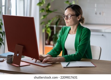 Successful Focused Caucasian Businesswoman Working On Computer Sits At Office Desk. Business Lady In Glasses And Green Jacket Is Typing Order For Company Employees Or E-mail Letter For Partners