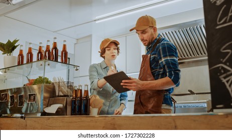 Successful Food Truck Man and Woman Employees in Brown Caps are Using a Tablet Computer. They Discuss Their Profits and Future Business Plans. Commercial Truck or Kiosk Selling Street Food and Drinks. - Powered by Shutterstock