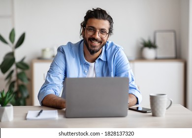 Successful Freelancer. Happy Millennial Indian Man Sitting At Desk With Laptop, Smiling And Posing At Camera While Working On Computer At Home Office, Wearing Eyeglasses And Braces, Free Space