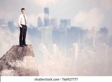 A Successful Good Looking Businessman Standing On Top Of A High Cliff Above The City Scape With Clouds Concept