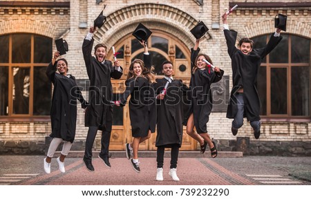 Successful graduates in academic dresses are holding diplomas, looking at camera and smiling while jumping for the photo outdoors