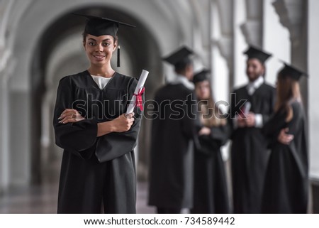 Successful graduates in academic dresses are talking in university hall, girl in the foreground is holding diploma, looking at camera and smiling