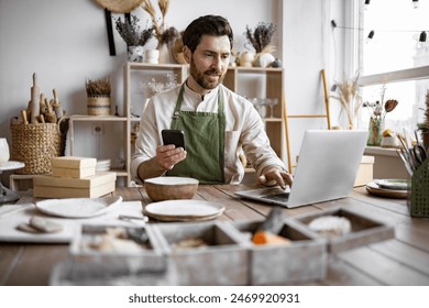 Successful guy store employee with ceramic tableware shoots photos on smartphone. Charismatic Caucasian man seller in souvenir shop takes pictures of goods at phone for posting on social networks. - Powered by Shutterstock