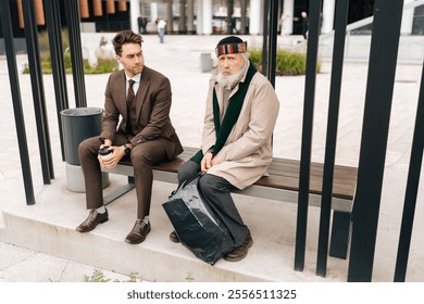 Successful handsome businessman in suit holding coffee cup, talking and listening to sad senior adult homeless man while sitting on urban bench at bus stop. Concept of helping older tramp people. - Powered by Shutterstock