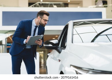 Successful Handsome Caucasian Shop Assistant In Formal Clothes Holding Clipboard Writing Checking Car Options Information At Automobile Dealer Shop Store.