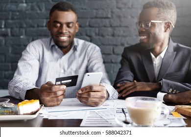 Successful Happy African-American Businessman Paying Bill With Credit Card Online Via Internet, Making Transaction, Using Mobile Bank Application On Cell Phone During Meeting With His Partner At Cafe