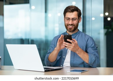 Successful and happy businessman with beard works in modern bright office, male boss uses laptop, business owner in casual shirt holds phone, browses internet pages - Powered by Shutterstock