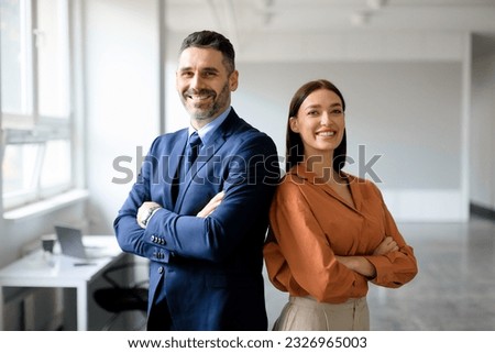 Successful happy businessman and businesswoman standing back to back with arms crossed and smiling at camera, office interior. Partnership concept