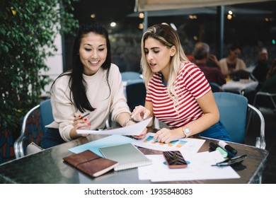 Successful hipster girls talking during cooperation meeting for checking informative documents in street cafe, diverse female planning organisation while doing university homework and course work