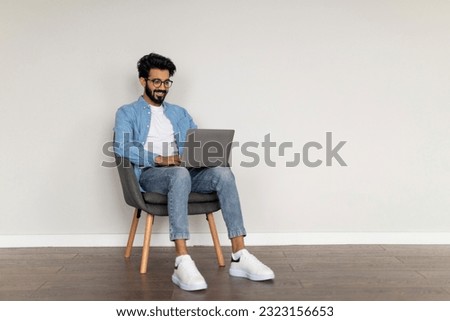 Successful Indian Freelancer Guy Using Laptop, Working Online While Sitting In Armchair Indoors, Eastern Millennial Man Browsing Internet On Computer Or Writing Article For Blog, Copy Space