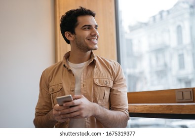 Successful Indian Man Using Smart Phone With Mobile App For Ordering Food Online On Website. Happy Asian Guy Holding Cellphone, Chatting, Receive Text Message, Booking Tickets, Looking At Window