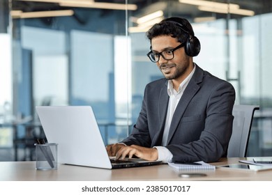 Successful indian programmer working inside office with laptop, man in headphones and business suit, typing code on keyboard, developing new software.