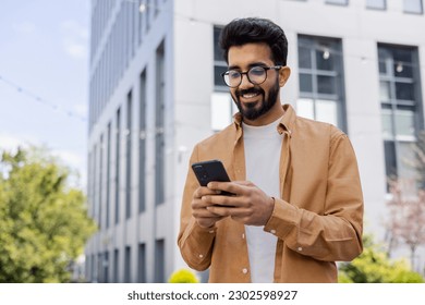 Successful Indian young businessman outside office building walking in daytime, man holding phone in hands, businessman dialing, browsing online pages, programmer engineer in glasses. - Powered by Shutterstock