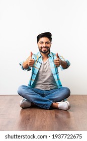 Successful Indian young man celebrating victory with thumbs up sign, sitting isolated over wooden floor against white background