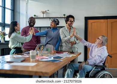 Successful Intercultural Colleagues Giving Each Other High Five As Symbol Of Team Building While Standing By Table With Financial Papers