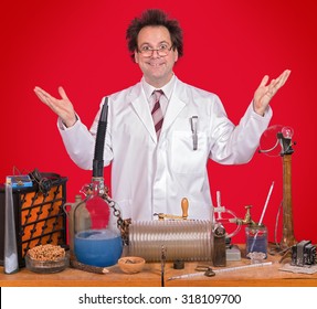 Successful Inventor At His Desk With Equipment On Red Background. Genius Professor At The Desk. Researcher In The Lab. Successful Research In The Science Lab. Mad Genius Shows His Hand Up Over Desk.