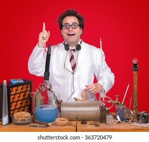 Successful Inventor At His Desk With Equipment On Red Background. Genius Professor Shows Triumph For Invention. Successful Scientific Research. The Inventor Of Celebrating Success In The Lab.