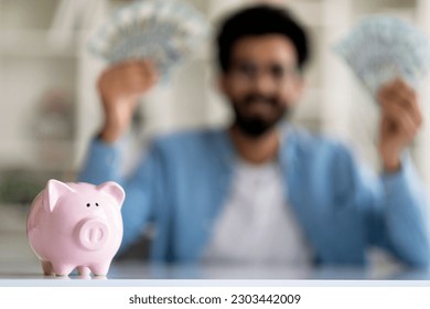 Successful Investment. Closeup Shot Of Piggy Bank Standing On Table With Happy Indian Man Holding Cash In Both Hands On Background, Lucky Eastern Male Demonstrating Profit, Selective Focus