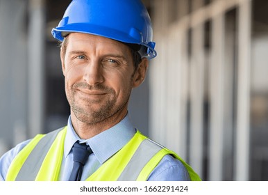 Successful Male Architect At Building Site Looking At Camera. Confident Construction Manager Wearing Blue Helmet And Yellow Safety Vest With Copy Space. Portrait Of Successful Mature Civil Engineer.