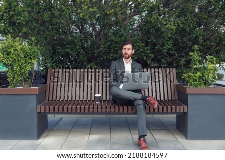 Successful male businessman working on laptop at lunchtime sitting on bench near office
