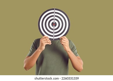 Successful male freelancer who in search of target audience covers his face with darts board. Unknown young man in casual T-shirt hides his face behind darts on khaki background. Targeting concept. - Shutterstock ID 2260331339