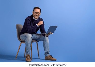 Successful male manager with laptop smiling at camera while sitting on chair over blue background - Powered by Shutterstock