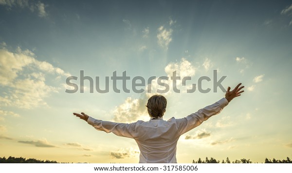 Successful\
man in elegant white shirt standing with his back to the camera\
with his arm spread widely towards beautiful majestic evening sky\
as he celebrates his success and\
prosperity.