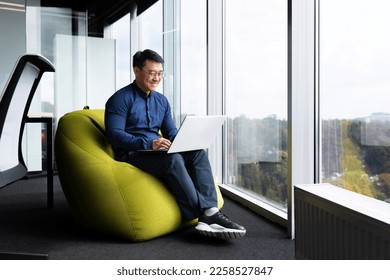 Successful mature asian working inside office using laptop, man typing on keyboard and smiling, businessman in shirt and glasses satisfied with work and achievement results, programmer at work. - Powered by Shutterstock