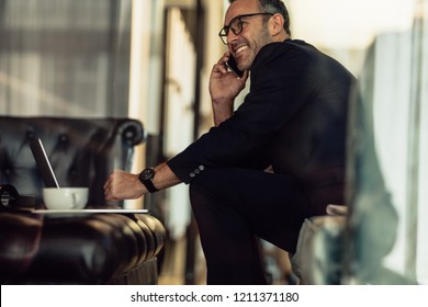 Successful Mature Businessman Sitting In Hotel Lobby Talking On Cellphone. Happy Man In Business Suit Working From Hotel Lobby.