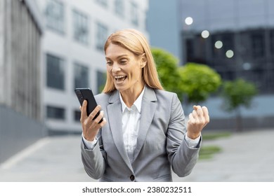 Successful mature joyful businesswoman received online notification message with good results achievement on phone, woman boss uses app on smartphone, walks outside office building in business suit. - Powered by Shutterstock