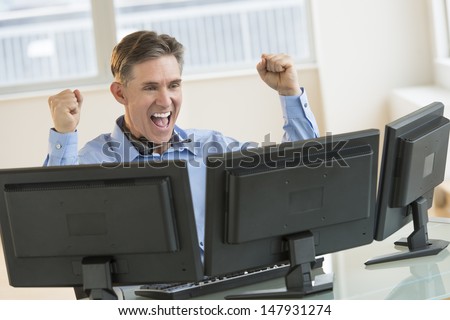 Successful mature male trader screaming while using multiple computers at desk in office